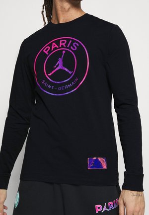 Mann trägt ein schwarzes Langarmshirt mit pinkem und lila Paris Saint-Germain-Logo und passende schwarze Hose mit Paris-Schriftzug und Emblem.