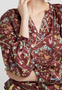 Blouse bourgogne à motif floral, fabriquée en tissu léger, avec un col en V profond, des manches froncées et des détails délicats au niveau du col et de l'ourlet.