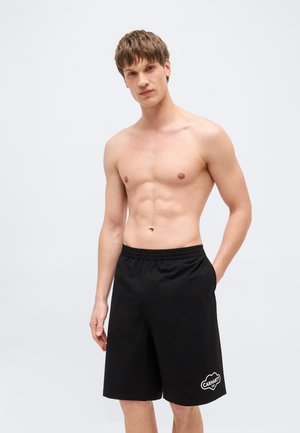 Uomo a torso nudo che indossa pantaloncini neri Carhartt larghi, in piedi con una mano in tasca davanti a uno sfondo semplice.