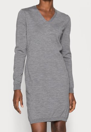 Kobieta ubrana w długą, szarą dzianinową sukienkę z dekoltem w serek oraz ściągaczami na mankietach i u dołu sukienki.