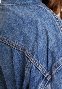 Denimjacka i en urblekt blå färg, med synliga sömmar, avslappnad passform och textil med struktur. Betonar ärm- och axelsömmarna.