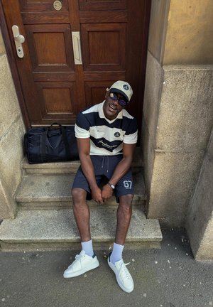 Άνδρας με καπέλο, γυαλιά ηλίου, ριγέ πουκάμισο, σορτς και λευκά αθλητικά καθισμένος σε πέτρινα σκαλιά δίπλα σε ξύλινη πόρτα, με μαύρη αθλητική τσάντα κοντά.