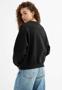 Sudadera negra con mangas raglán, con cuello de barco y un sutil detalle en el parche trasero, combinada con jeans de un azul claro.
