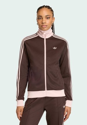 Femme aux cheveux tressés portant une veste de survêtement Adidas marron et rose clair, assortie à un pantalon, debout devant un fond uni.