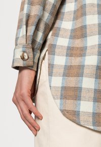 Hand rustend naast beige broek, gekleed in een blauw en beige geruit shirt met knoopmanchetten en een afgeronde zoom.