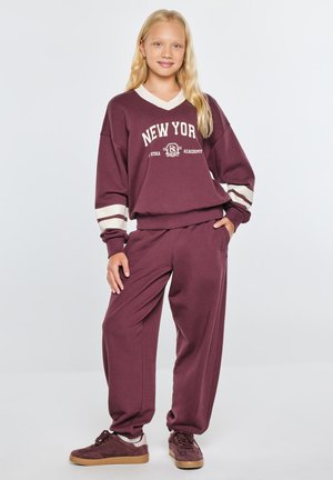 Młoda dziewczyna o długich blond włosach, ubrana w bordową bluzę New York i pasujące spodnie oraz brązowe trampki, stoi na jednolitym tle.