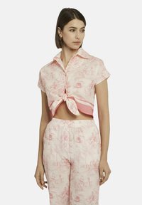 Kurzärmliges, rosa Hemd mit Kragen und einem geknoteten Front, das ein detailliertes Toile-Muster aufweist. Kombiniert mit passenden Hosen, die einen stimmigen Look erzeugen.