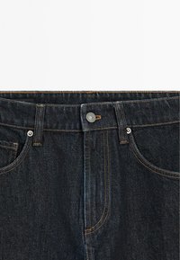 Mørkeblå denimjeans har et klassisk fem-for-lomme design, sølvknaplukning og kontrasterende tan syninger langs sømmene.
