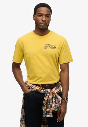 T-shirt giallo a maniche corte con una stampa grafica vicino al petto. Una camicia a quadri è legata attorno alla vita. Sono visibili jeans scuri e un orologio.