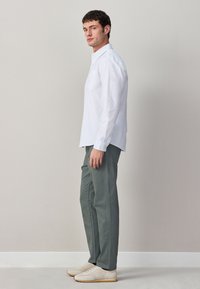 Jeune homme debout de profil portant une chemise blanche à boutons, un pantalon gris et des baskets beiges sur un fond uni gris clair.