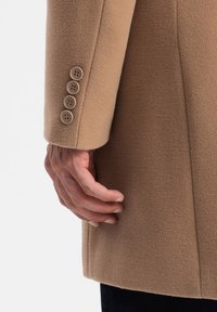 Kamelullskappa med ärm som visar en mjuk textur, fyra ljusbruna knoppar och en figursydd design. Handen vilar mot kappan.