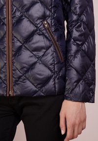 Marint quiltad jacka med diamantmönster, med en brun dragkedja och ficka, tillverkad av glänsande, lätt material.