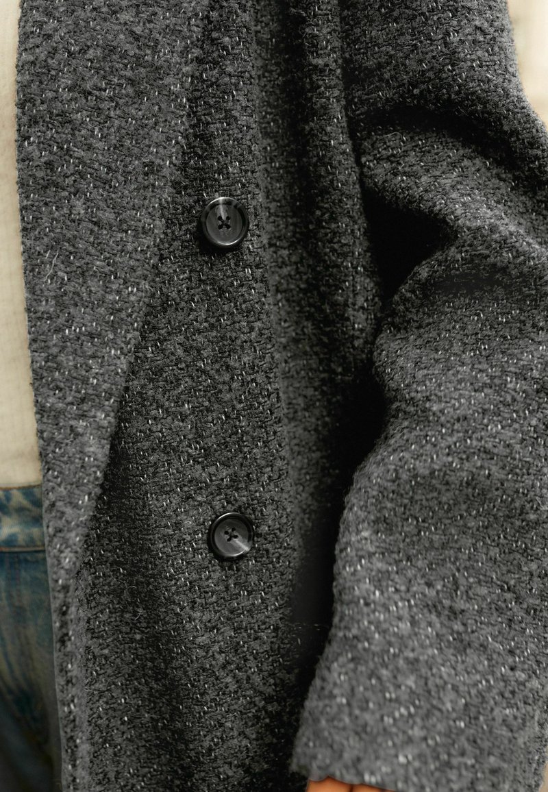 Cardigan texturé gris foncé avec une coupe décontractée, doté de deux boutons noirs et d'un tissu tissé doux. Détails visibles aux poignets et au col.