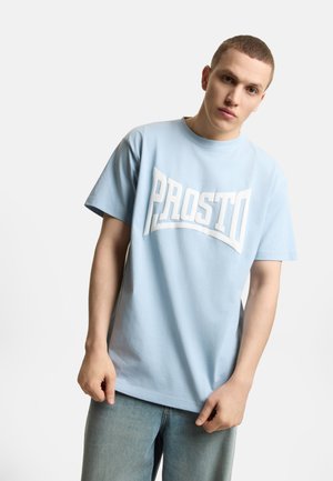 Młody mężczyzna w jasnoniebieskiej koszulce "PROSTO" i dżinsach, stoi na tle jednolitego jasnego tła, patrząc lekko w dół.