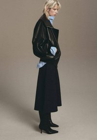 Chaqueta oversize de cuero negra, camisa de rayas azul clara debajo, falda midi negra y botas de tacón puntiagudas negras. Fondo sencillo.