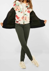 Giacca bomber floreale con base crema, caratterizzata da fiori rosa e rossi, sovrapposta a un top verde e pantaloni skinny oliva, abbinata a sneakers beige.