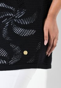 Schwarzes gestricktes Oberteil mit ausgeschnittenen Blumenmustern, strukturierten Streifen und einem goldenen Akzentknopf nahe dem Saum. Kombiniert mit weißen Hosen.