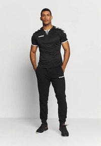 Camiseta deportiva negra con detalles y logo blancos, combinada con pantalones de chándal negros y zapatillas negras, posando frente a un fondo neutro.