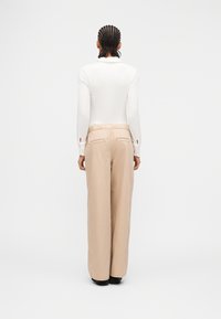 Άτομο  με πλεγμένα μαλλιά στέκεται με την πλάτη, φοράει λευκή μπλούζα με μακριά μανίκια και μπεζ παντελόνι φαρδιάς γραμμής με πίσω τσέπες.