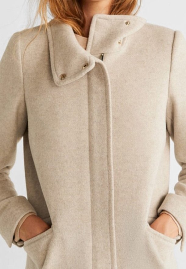 Manteau beige en mélange de laine avec col haut, fermeture à pression et poches latérales. Texture lisse et coupe ajustée renforcent son design moderne.