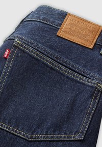 Jeans de mezclilla azul oscuro con un parche de cuero con texto en relieve, costuras amarillas y una etiqueta roja en el bolsillo trasero.