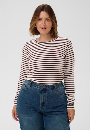 Femme portant un t-shirt à manches longues rayé blanc et rouge foncé avec un petit cœur rose sur la poitrine et un jean bleu taille haute, debout sur un fond blanc.