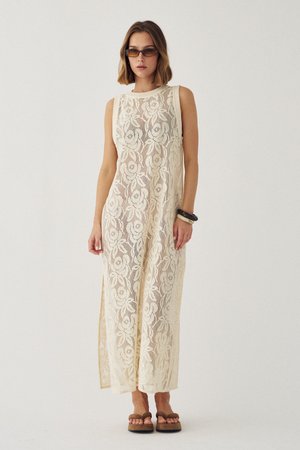 Žena v bezrukávových dlhých šatách krémovej farby z čipky s kvetinovými vzormi, slnečné okuliare, robustné sandále a náramky na zápästí, na jednoliatom pozadí.
