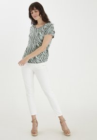 Zaļš un melns zebras raksta īso piedurkņu tops apvienots ar baltām slim-fit biksēm un bēšām siksnām uz platformas sandālēm. Visā apģērba virsmā ir gludas tekstūras.