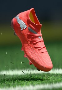 Fényes narancssárga futballcipő texturált mintával, kontúrozott kialakítással és fekete logóval. Cipőfűzőkkel és alatta megmozduló fűvel.