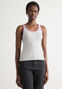 Žena u sivoj majici bez rukava s crvenim detaljima i logom Calvin Klein, uparena s crnim hlačama, stoji protiv jednostavne bijele pozadine.