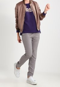 Chaqueta bomber marrón claro con puños acanalados, superpuesta a un sudadera púrpura con "CRE" en color beige. Pantalones grises ajustados y zapatillas blancas.