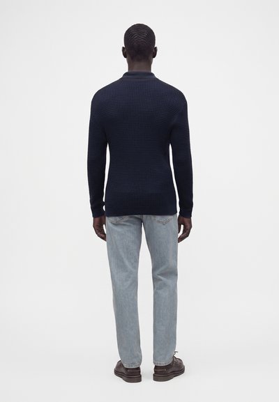 Homme debout, dos tourné, portant un pull texturé foncé, un jean bleu clair et des chaussures marron foncé sur un fond blanc uni.