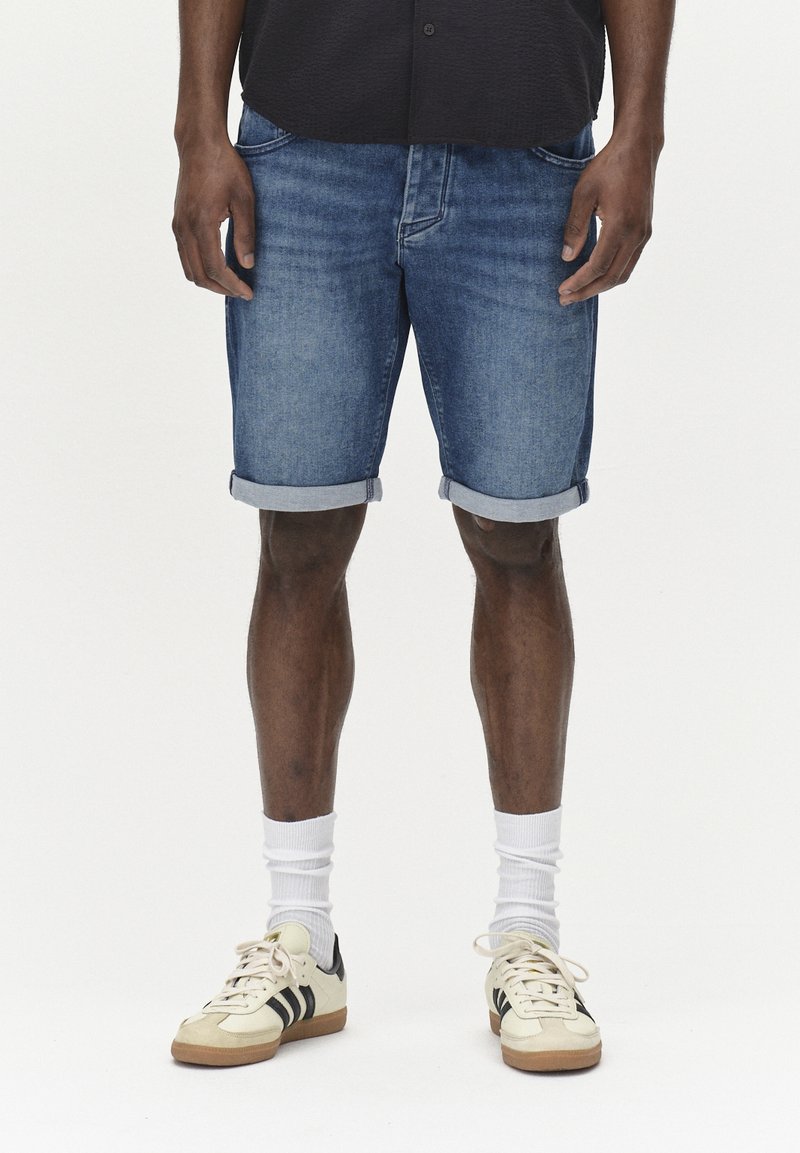Pantalones cortos de mezclilla en azul claro con dobladillos enrollados, combinados con calcetines blancos y zapatillas crema con franjas negras. Diseño simple y casual.