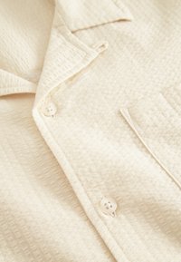 Chemise couleur crème avec un motif texturé, dotée d'un col cranté, de deux boutons et d'une poche poitrine. Tissu doux avec une légère brillance.