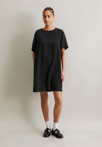 Black, loose-fitting T-shirt dress with short sleeves, round neckline, and a smooth texture. Worn with black shoes and white socks.