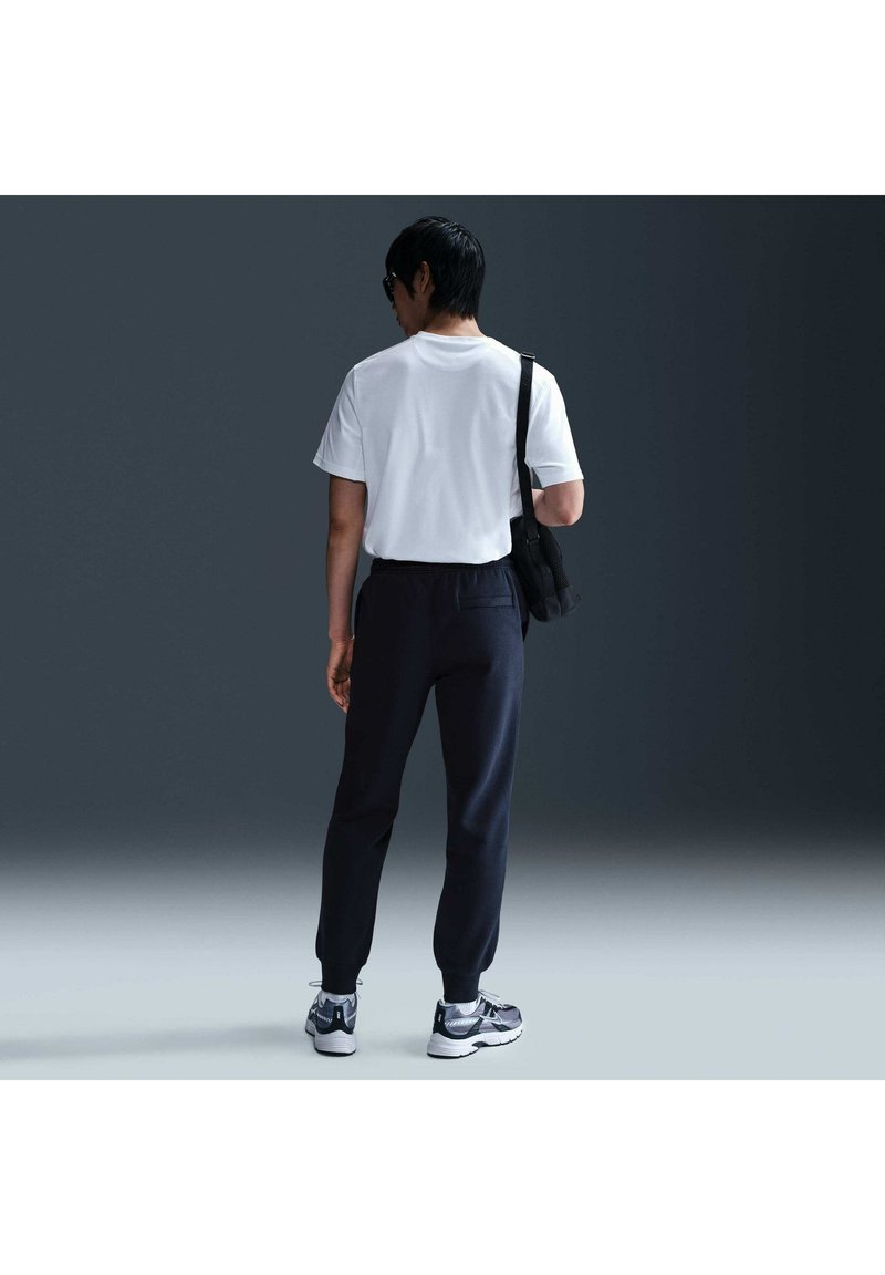 Persona di spalle, con t-shirt bianca, pantaloni neri, sneakers, occhiali da sole e borsa nera a tracolla, in piedi su uno sfondo scuro.