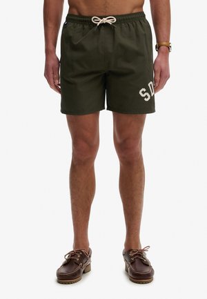 Mann trägt dunkelgrüne Shorts mit Kordelzug und "SD"-Beschriftung am linken Bein, braune Leder-Bootsschuhe und eine braune Armbanduhr am rechten Handgelenk.