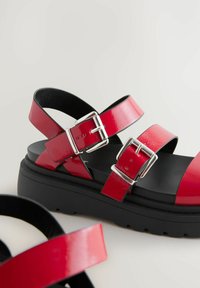 Sandales en cuir verni rouge avec deux brides réglables à boucles, une semelle plate noire et un pied de mémoire en texture lisse.