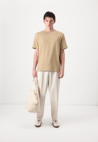 Beige Baumwoll-T-Shirt, lockere Passform, kombiniert mit cremefarbenen Hosen. Das Model hält eine weiche, neutralfarbene Tragetasche mit Schulterriemen.