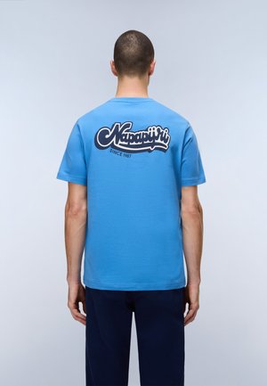 Uomo che indossa una T-shirt blu Napapijri con logo e scritta "Since 1987" sul retro, in piedi davanti a uno sfondo chiaro e uniforme.