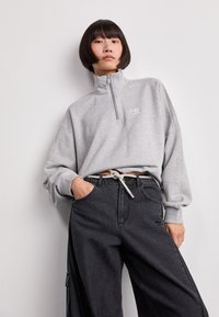 Sudadera gris con cremallera y alto cuello, con un pequeño logo. Combinada con pantalones cargo oscuros, que destacan un detalle de cordón metálico.
