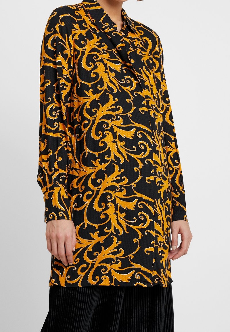 Chemise noire à manches longues avec un motif floral orné orange vif portée par une personne avec la main reposant sur le côté.
