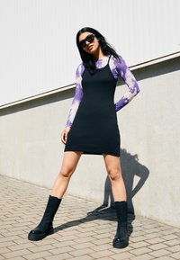 Svart ribbad miniklänning i kombination med en lila tie-dye långärmad skjorta och kraftiga svarta knähöga stövlar. Släta texturer och figurnära design.