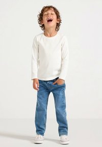 Valge pikaaegadega särk ümarate kaelusega, millele on lisatud sinised teksapüksid. Poiss seisab käed taskutes ja naeratab laialt.