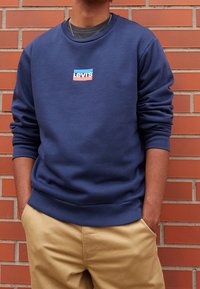 Sudadera azul marino con cuello redondo y un pequeño logo multicolor de Levi's en el pecho. Tela suave, puños y cinturilla acanalados. Pantalones de color beige visibles.