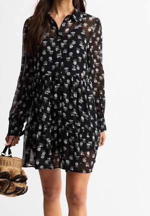 Donna che indossa un vestito nero trasparente a maniche lunghe con stampa floreale bianca, che tiene una borsa di paglia intrecciata con dettagli neri.