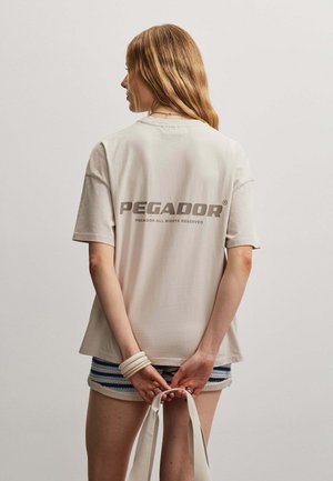 Frau mit langen Haaren steht mit dem Rücken zur Kamera, trägt ein beiges Pegador T-Shirt und gestreifte Shorts und hält weiße Taschenriemen hinter dem Rücken.