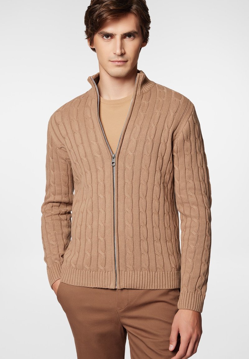 Beżowy sweter na zamek z wzorem w warkocze, ściągaczami przy rękawach i u dołu oraz gładką teksturą. Noszony na beżowej koszuli.