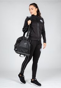 Conjunto deportivo negro con chaqueta con cremallera y pantalones ajustados con acentos blancos. La modelo sostiene un bolso de deporte negro con paneles de malla y logo.