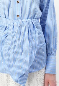 Blåstripet bluse med perleutstyr, en knyttet midje detalj og knappelukking. Lange ermer har stripete mansjetter og detaljer.